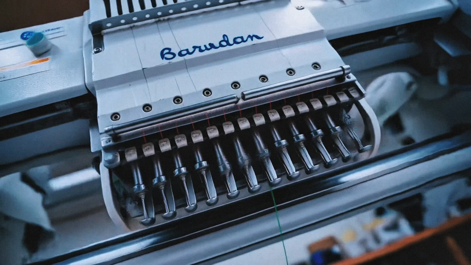 La devanture Maison Kouoh, des fils colorés et une machine à broder. Des plans rapprochés montrent la broderie d’un vêtement. Le film comprend des sous-titres intégrés à l’image.
